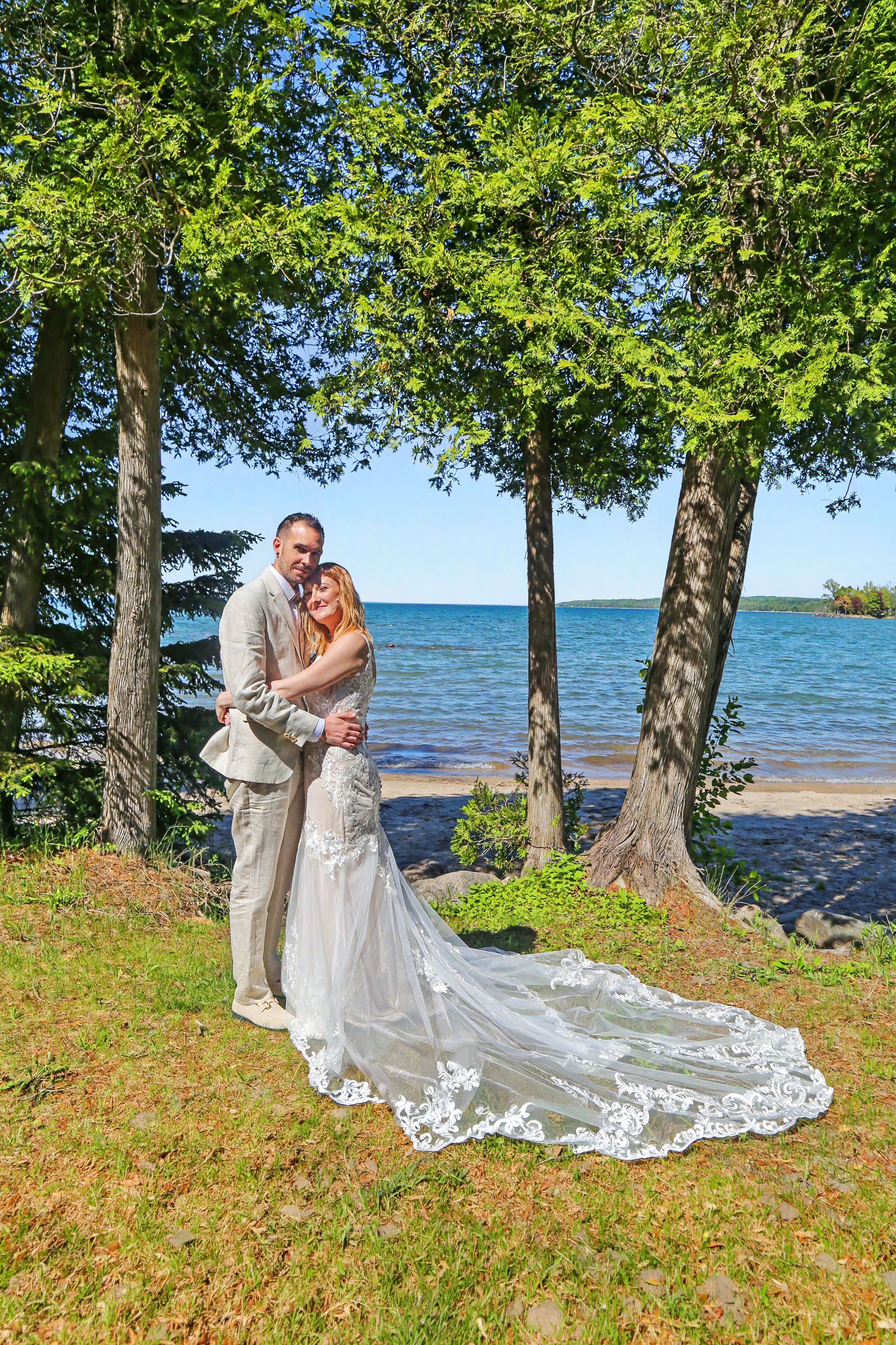 Bride and groom embracing near a lakeside with tall trees and blue water in the background on their wedding day.