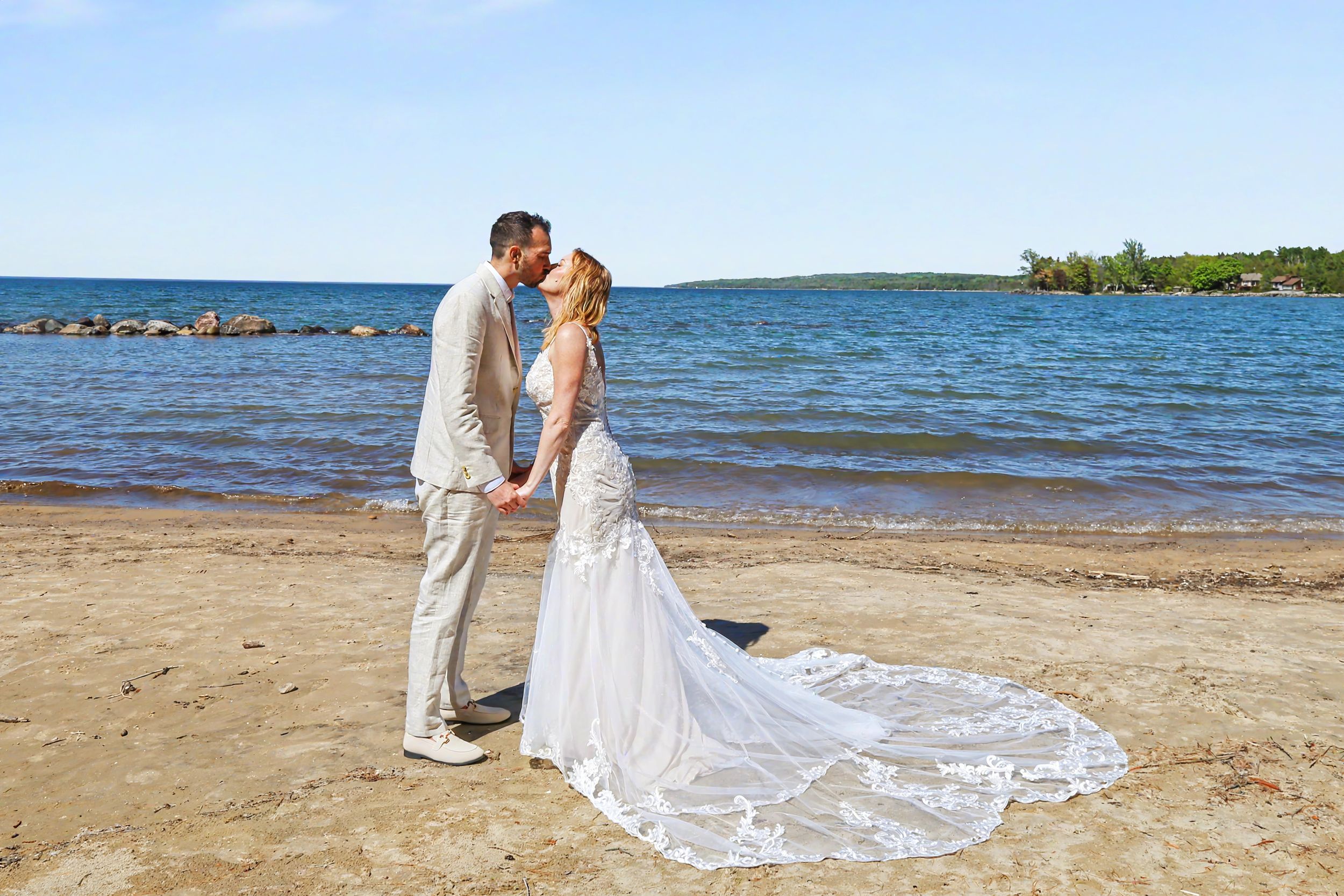 Newlywed couple sharing a kiss on a sandy beach, bride in white gown with long train, groom in tan suit.