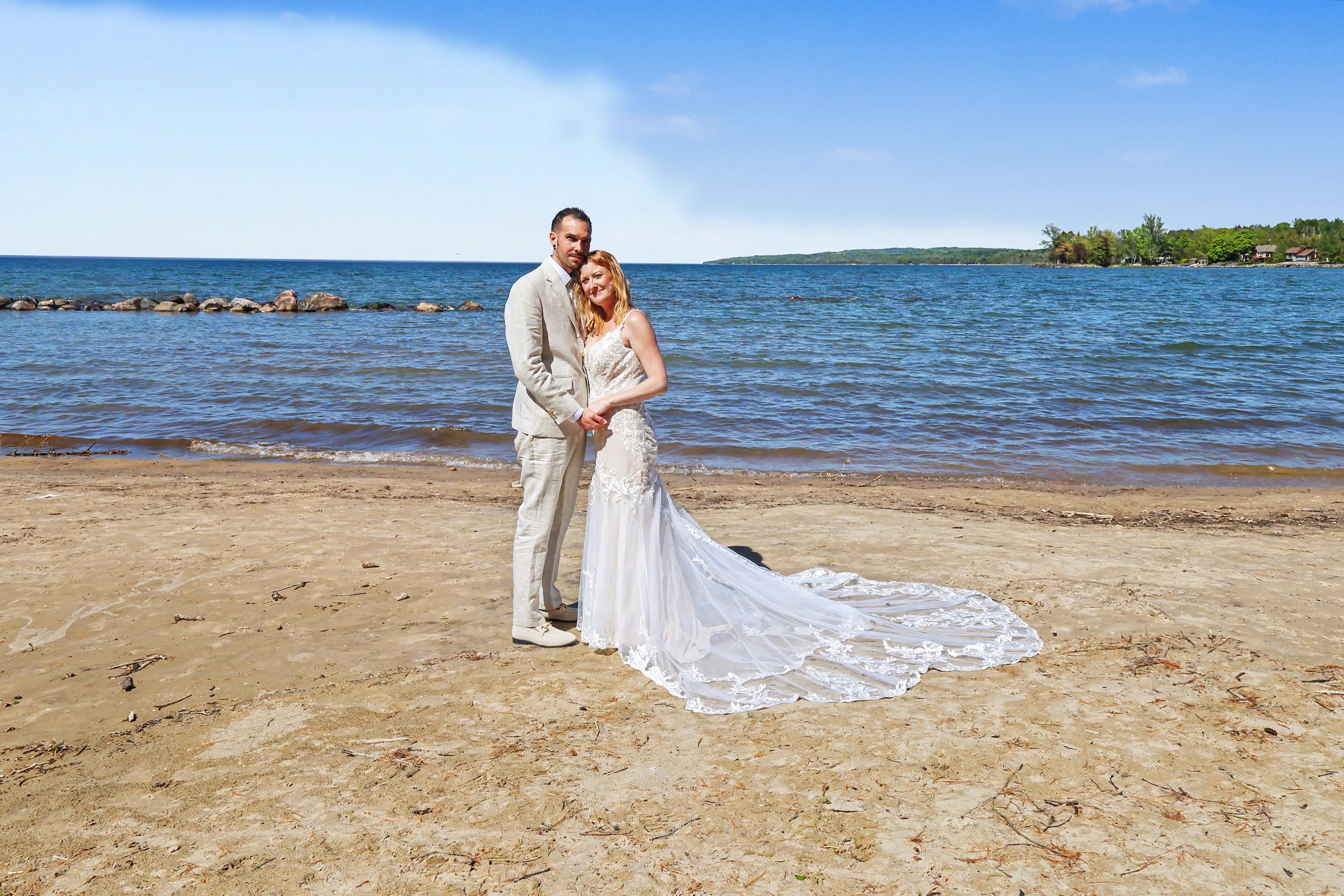 Bride and groom posing on sandy beach by lake, bride in lace gown with train, groom in tan suit.