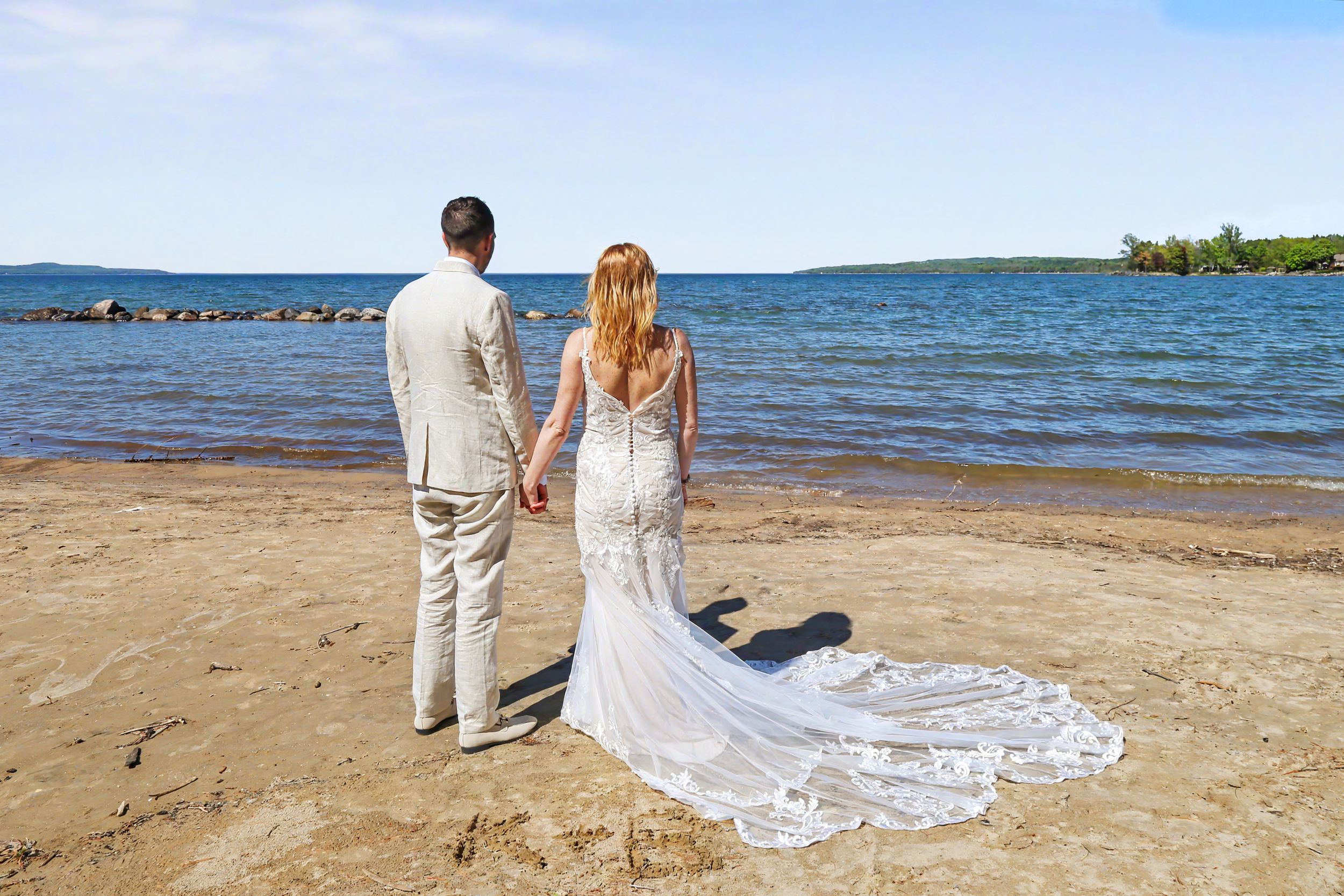 Bride and groom holding hands on sandy beach shore, woman in lace wedding gown with long train, sunny day.