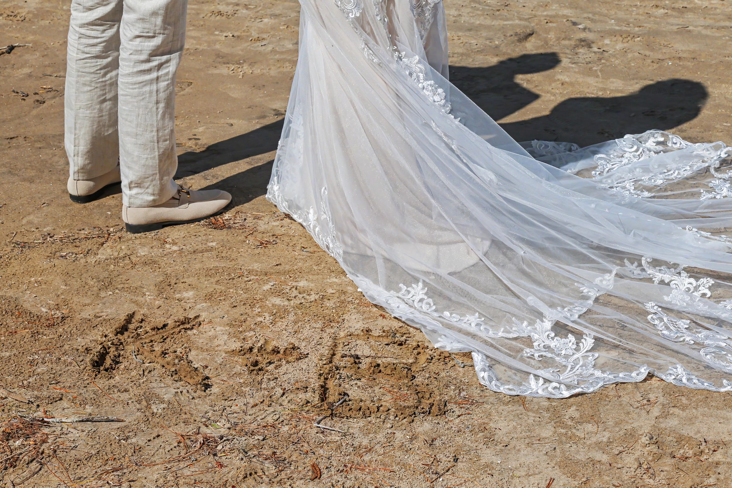 Bride in lace wedding gown with flowing veil train on sandy beach beside groom in beige suit.