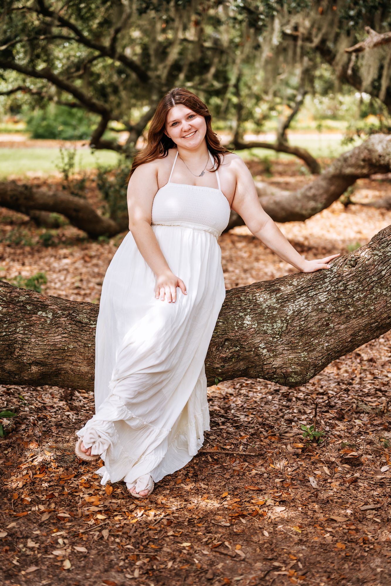 Young woman in white maxi dress posing gracefully on a large tree branch in a lush green park setting.