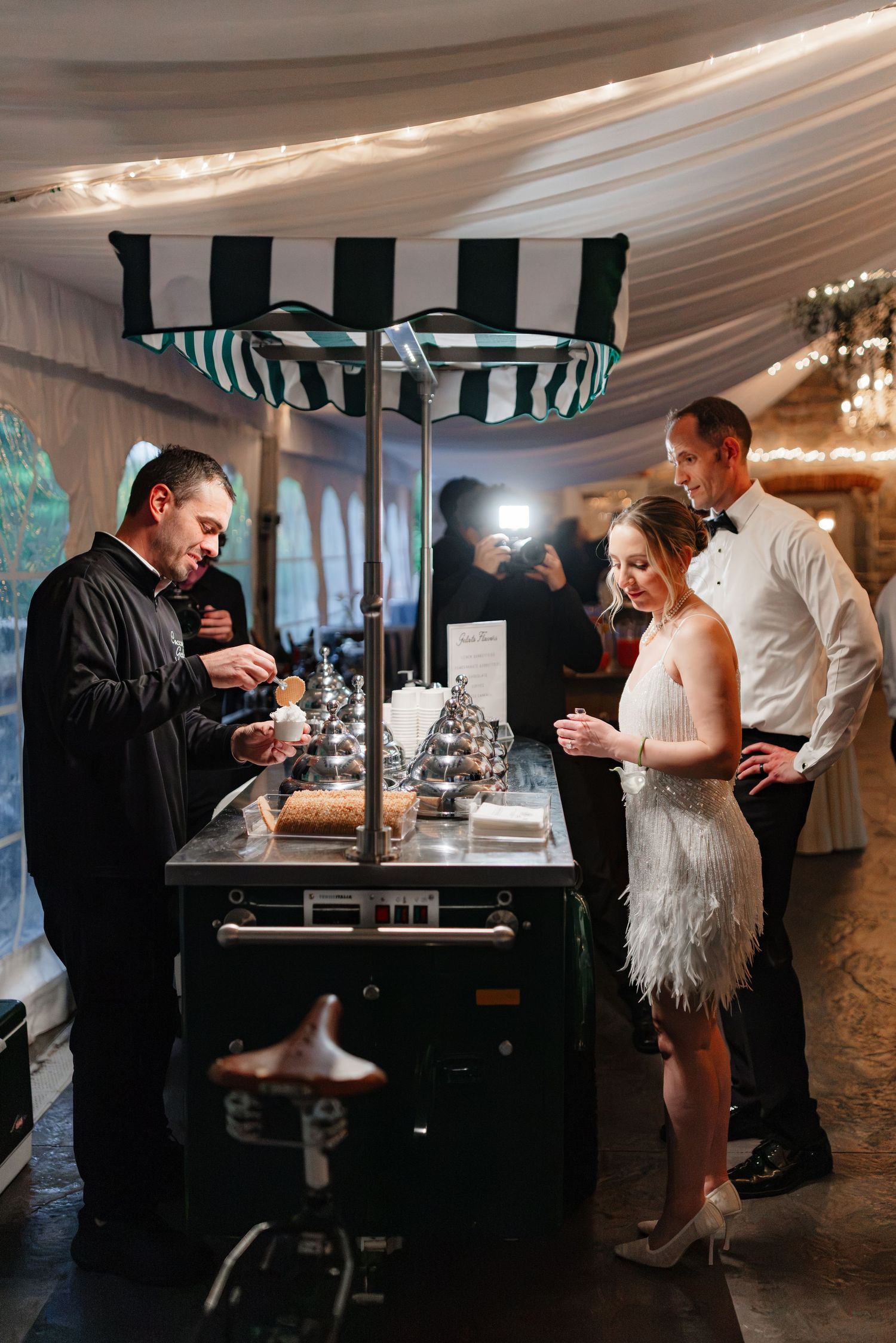 Bride in white fringe dress and groom ordering gelato from a striped cart at wedding reception.