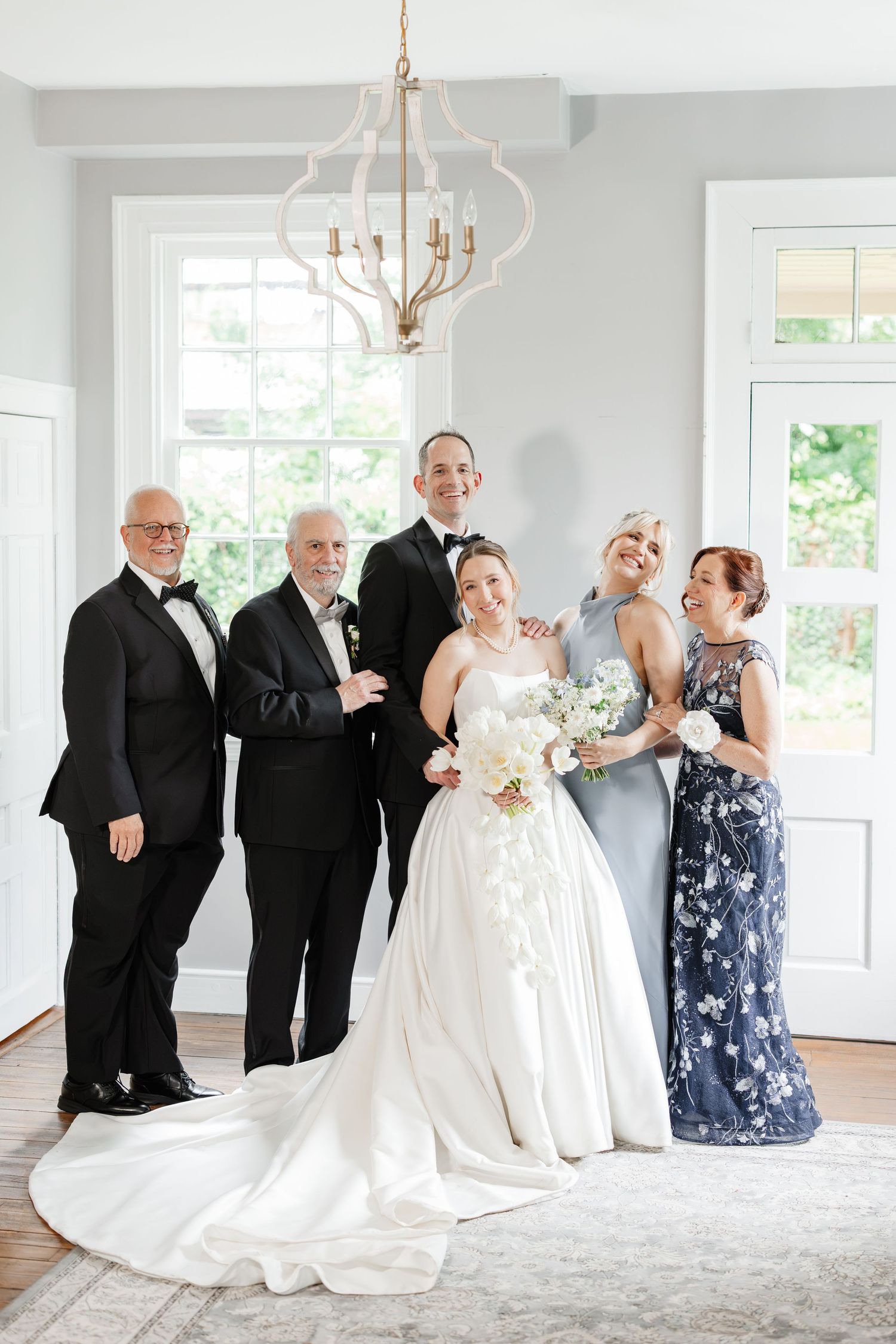 Wedding party group portrait with bride in white ball gown, groom in tuxedo, and family members in formal attire.