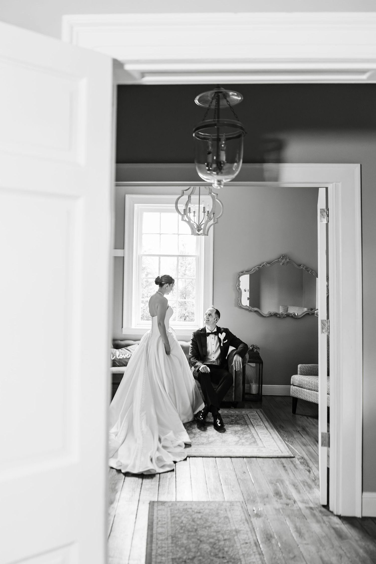 Black and white photo of bride in ball gown standing as groom sits in a chair in an elegant room.