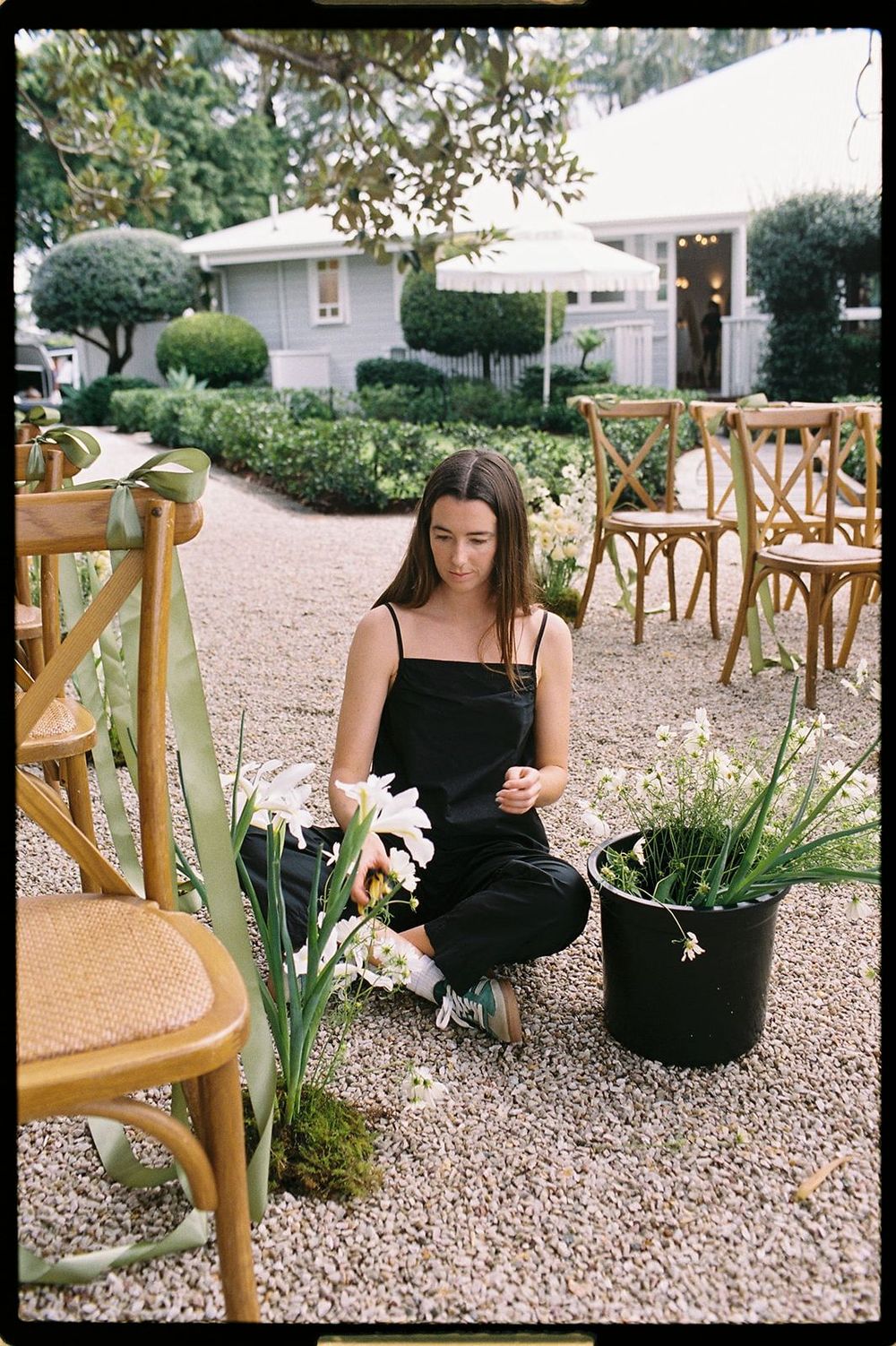Woman in black dress arranging white flowers outdoors at an elegant garden venue with wooden chairs on gravel path.