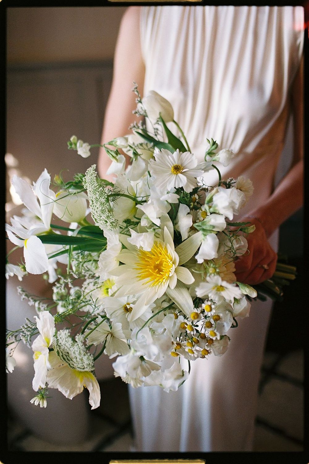 Bride holding a lush white and yellow wildflower bouquet with daisies, irises, and greenery in a romantic garden style.