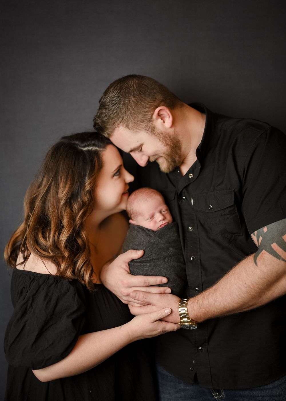 New parents tenderly hold their swaddled newborn baby together against a dark gray studio background.