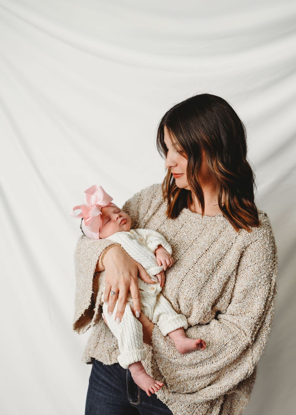 A young mother with dark hair holds her newborn baby wearing a pink bow against a white backdrop.