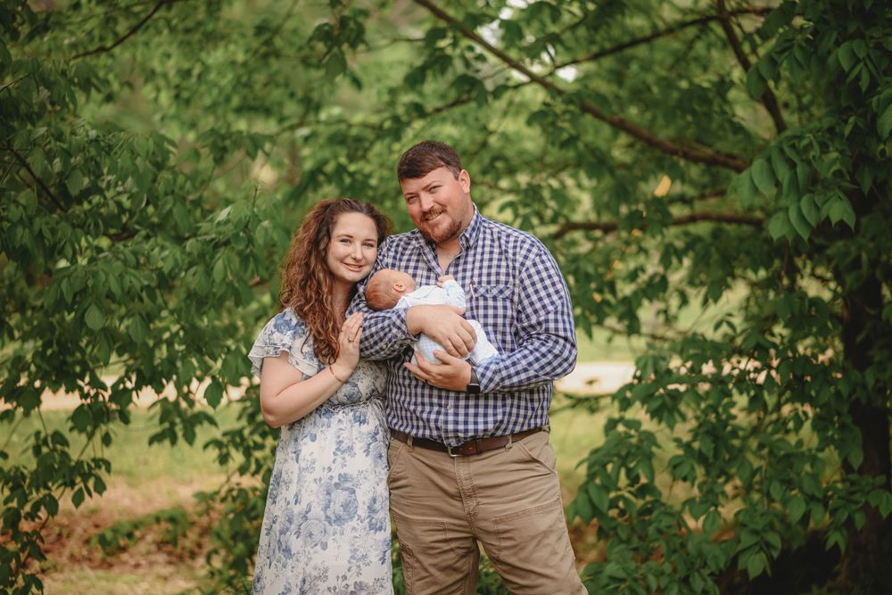 A couple stands outdoors under green trees, the man holding a newborn baby, both smiling in a lush garden setting.