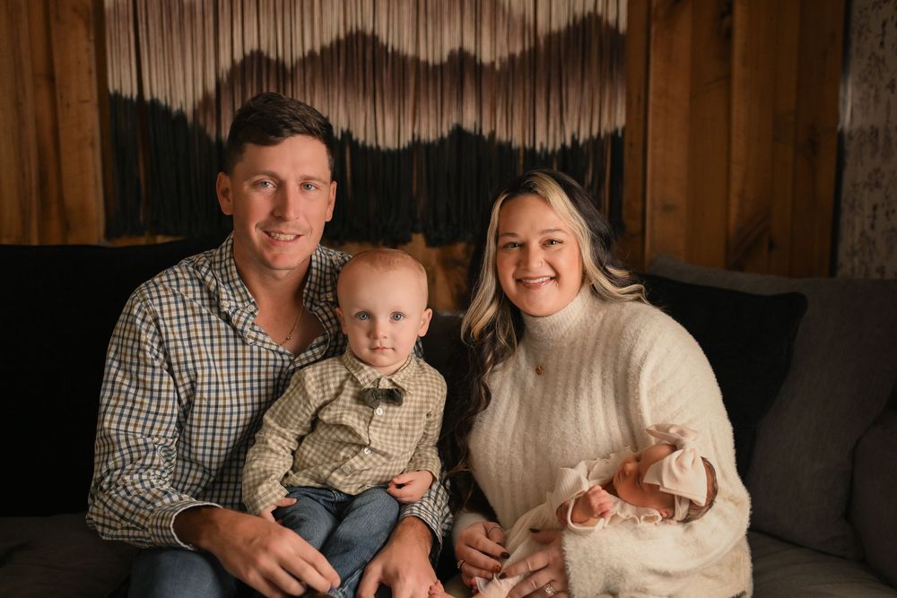 A smiling family of four poses on a couch, parents with a toddler boy in a bow tie and a newborn baby.