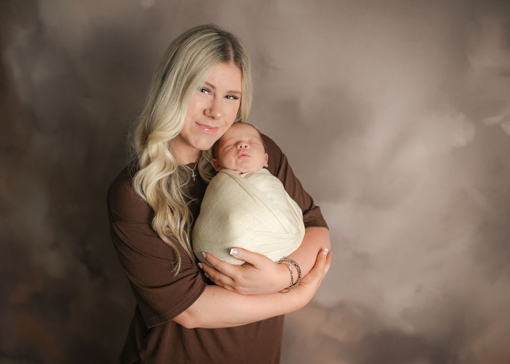 A mother in a brown dress tenderly holds her swaddled newborn baby against a warm taupe studio backdrop.