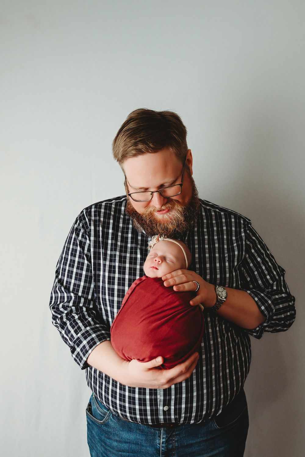 Bearded father in plaid shirt cradling sleeping newborn wrapped in red cloth, tender parenting moment.