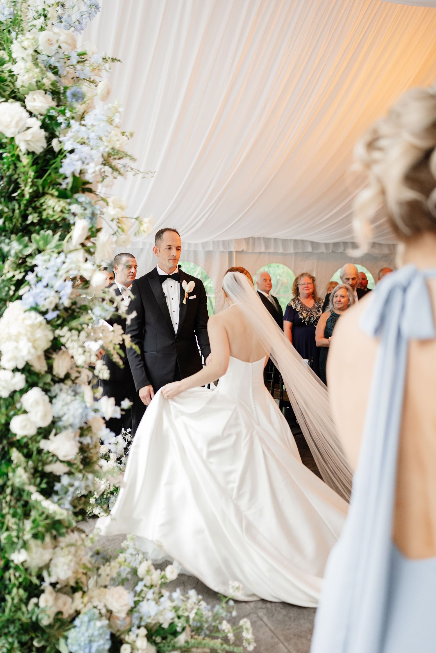 Bride in white gown with veil walks toward groom in tuxedo at floral wedding ceremony altar.