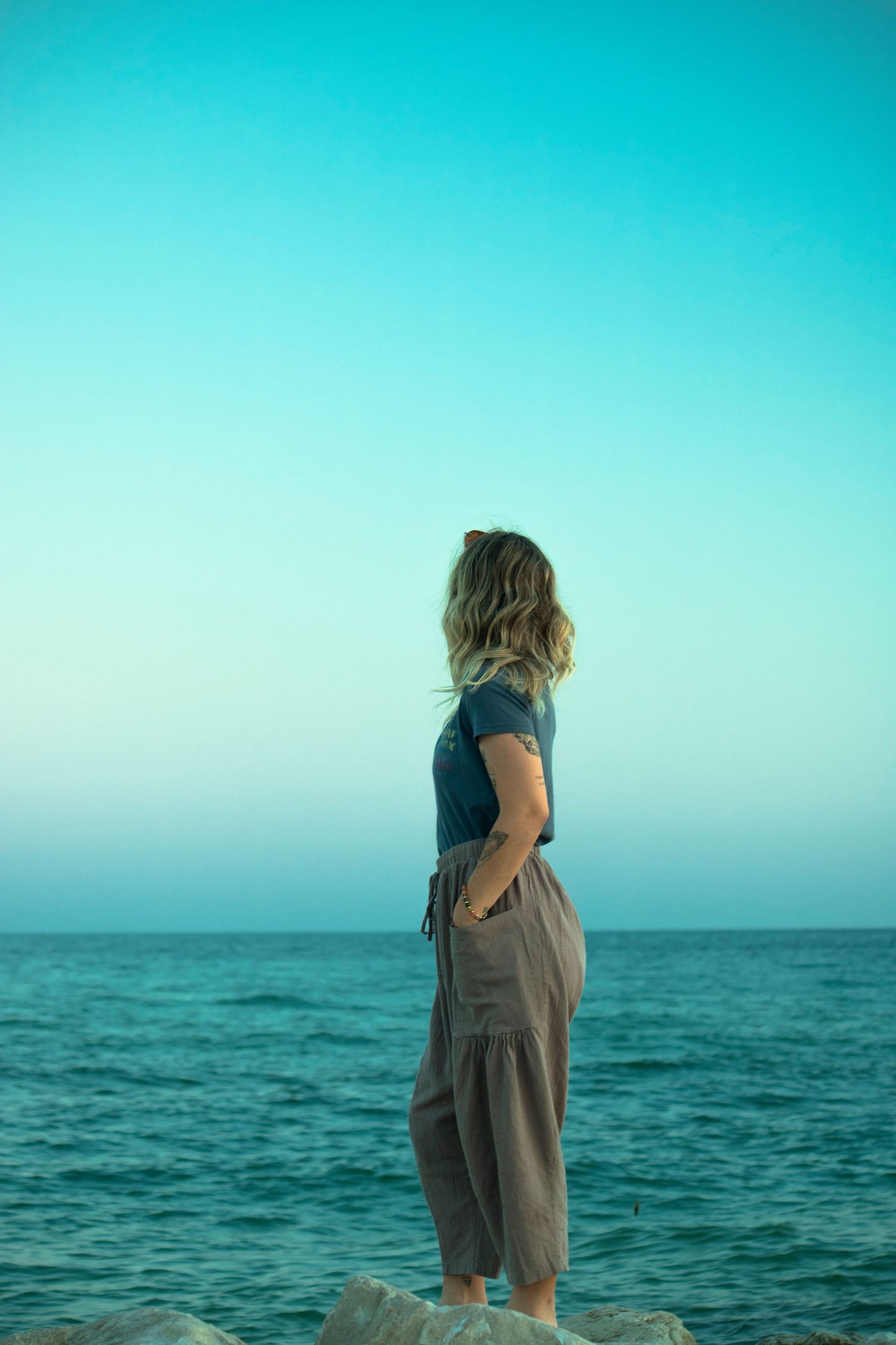 Woman standing on rocks gazing at turquoise ocean under clear blue sky, wearing gray pants and navy top.