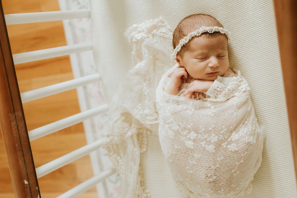 Newborn baby wrapped in white lace swaddle with floral headband sleeping peacefully in a crib.