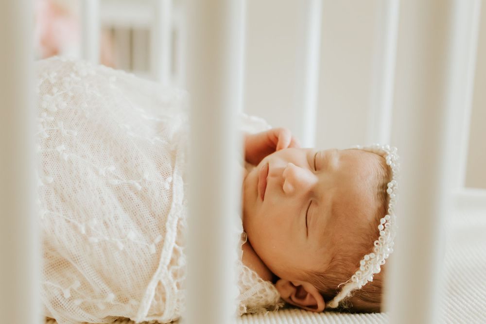 Peaceful newborn baby sleeping in a white crib, wrapped in a delicate lace blanket and bonnet.