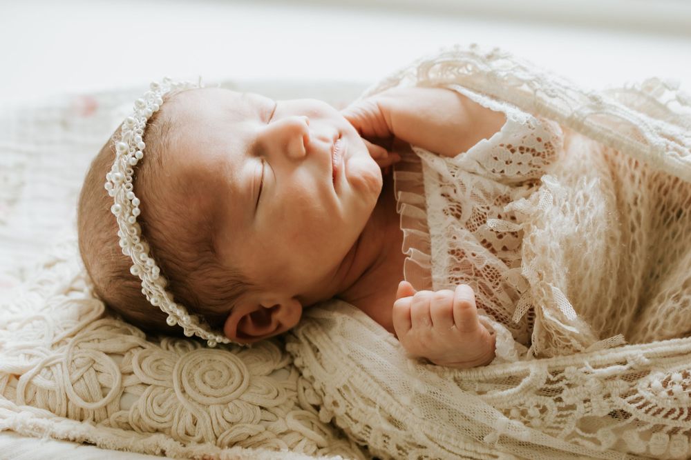 Peaceful newborn baby sleeping wrapped in delicate lace fabric with a floral headband on a cream blanket.