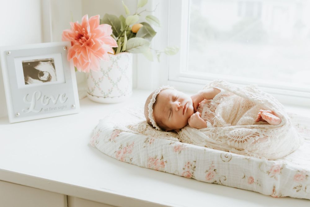 Newborn baby sleeping peacefully on floral changing pad near window with love frame and pink flowers.