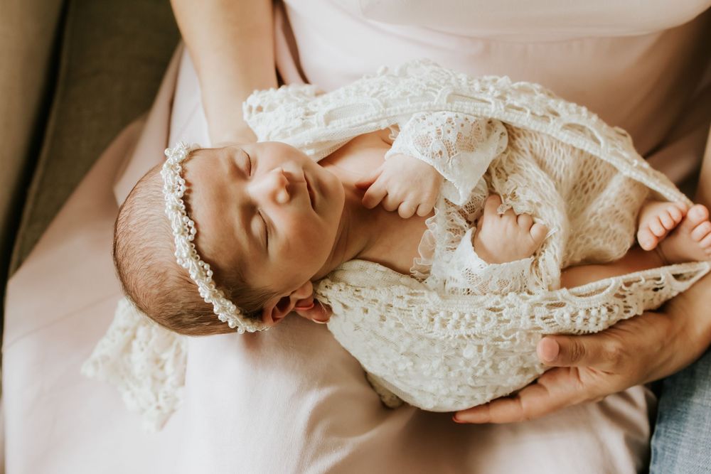 Newborn baby in white lace outfit and floral headband cradled in mother's arms.