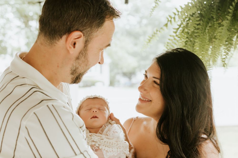 A couple smiles lovingly at their newborn baby wrapped in a white blanket outdoors.