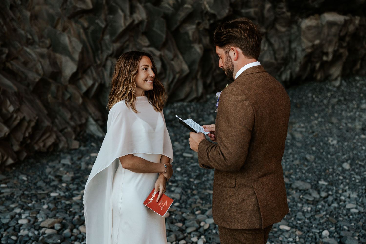 groom reading vows to bride while bride smiles with love in a cave/alcove at Reynisfjara Beach in Iceland during their elopement ceremony