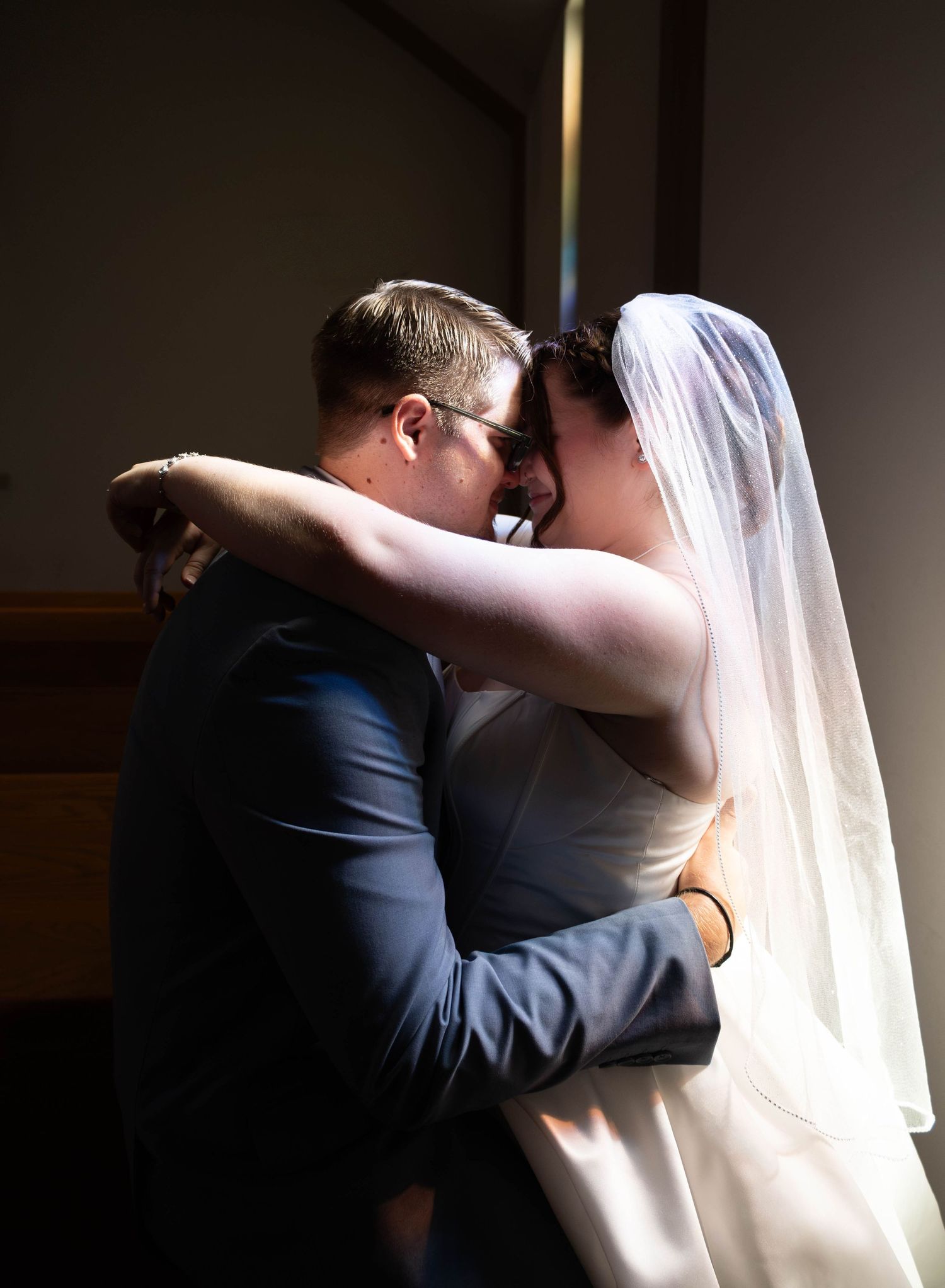 Bride in elegant white gown sitting on dark leather chesterfield sofa in moody, dimly lit room.