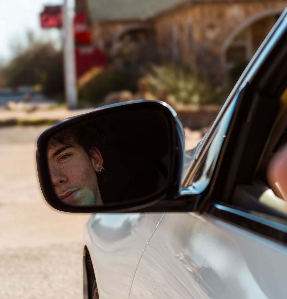 Young man with ear piercing reflected in a car side mirror outdoors.