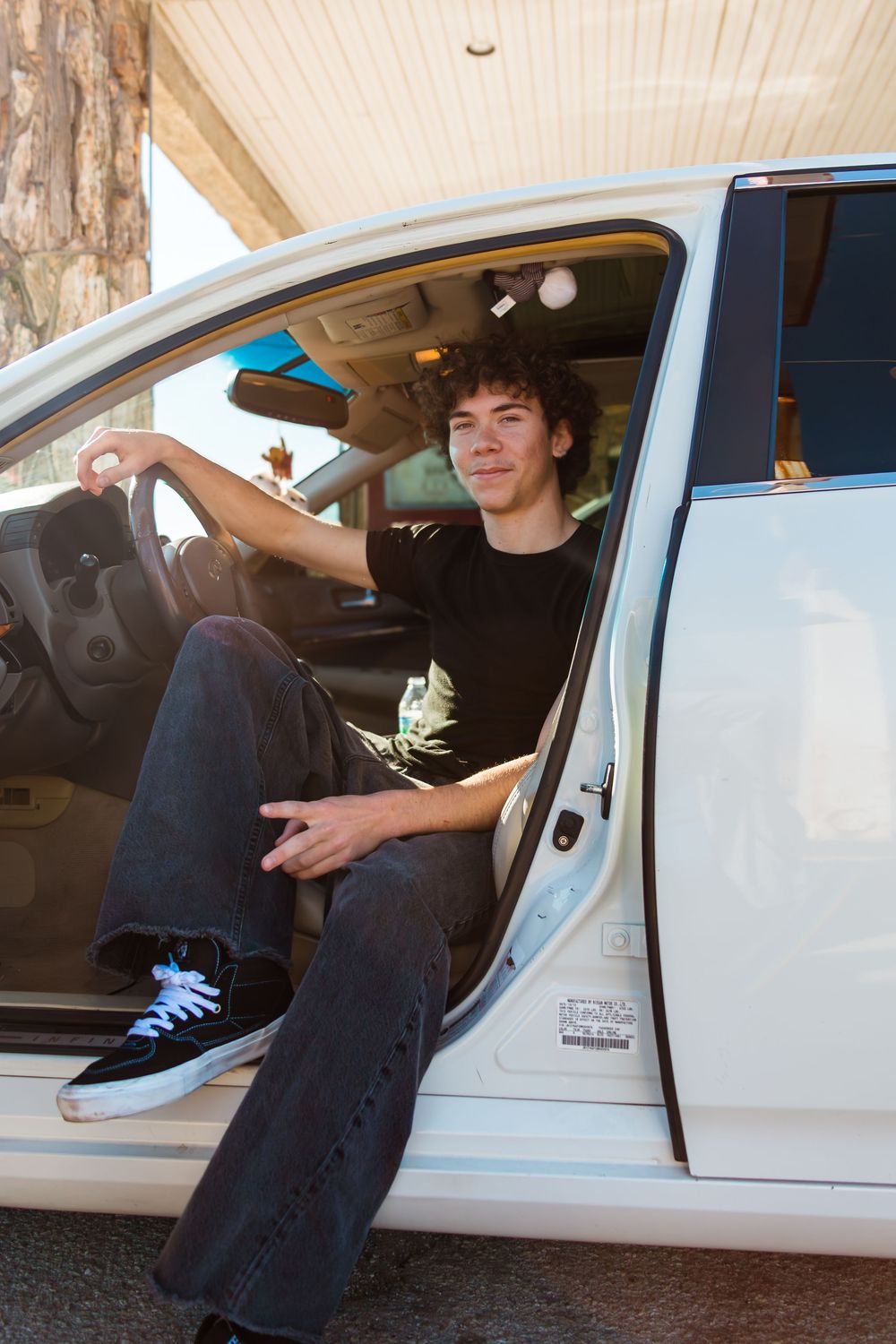 Young man with curly hair sitting in open car door, wearing black t-shirt and jeans, smiling casually.