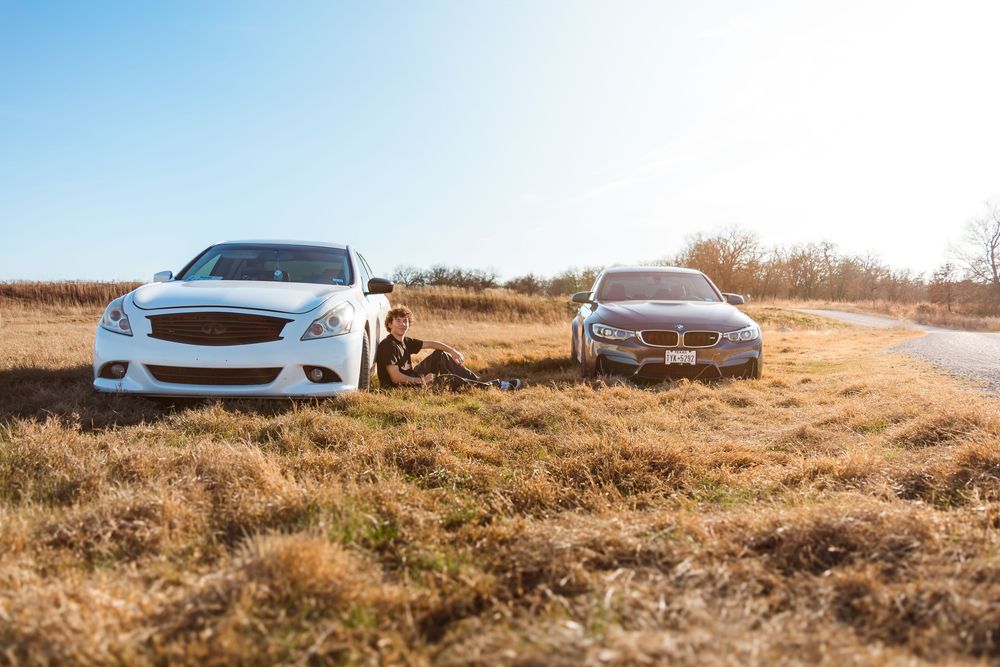 Two luxury cars, a white Infiniti and silver BMW, parked on dry grass field beside a rural road on a sunny day.