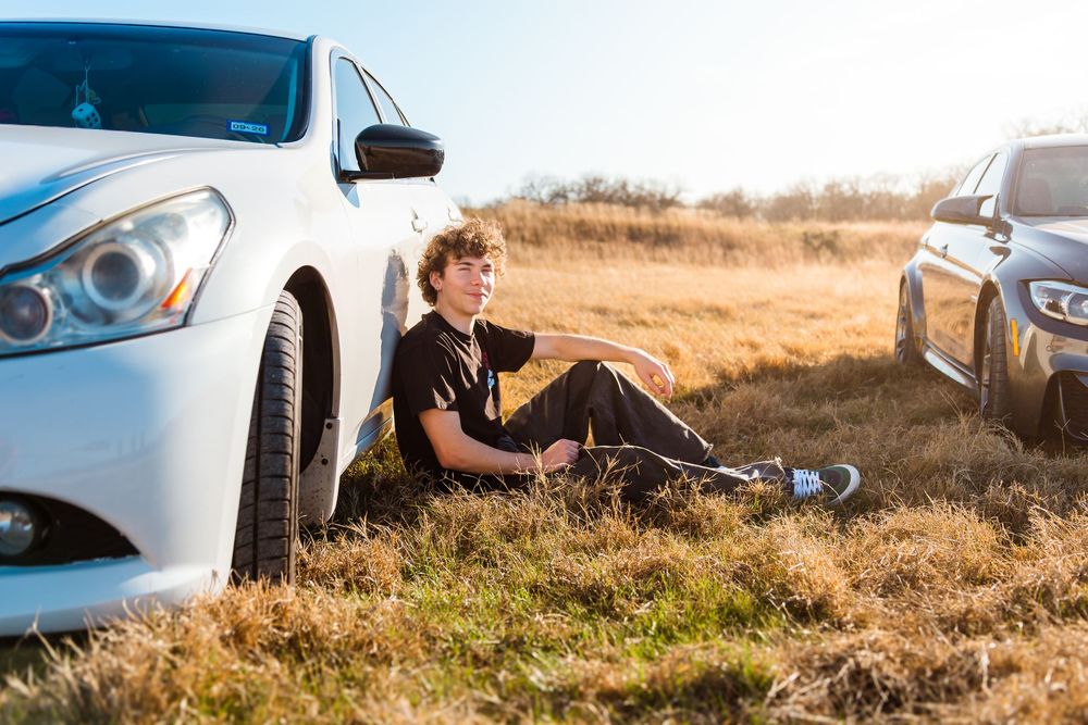 Young man sitting on grass between two cars in a sunny field, looking thoughtfully into the distance.