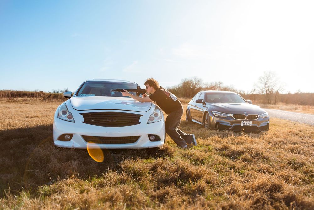 Man posing dramatically beside a white Infiniti and BMW on a sunny rural roadside with golden grass fields.