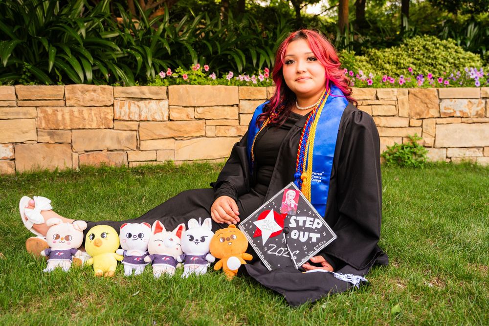 Graduate in black gown and blue sash sits on grass with plush toys and a decorated cap reading Stepped Out.