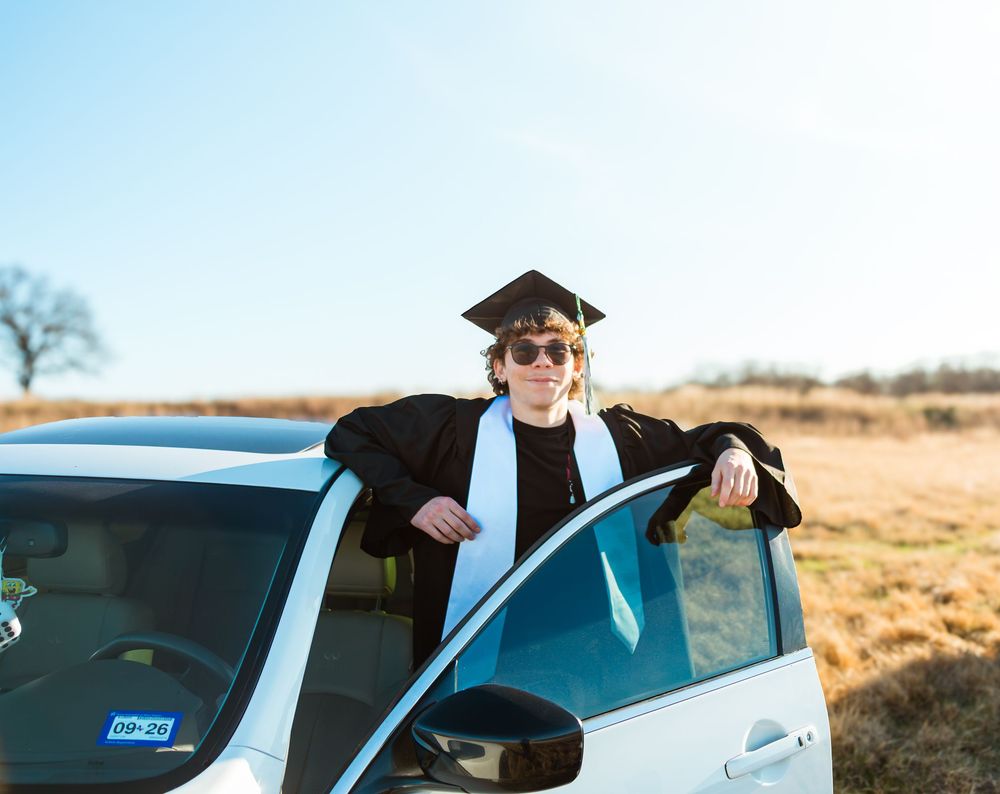 Young graduate in cap and gown leaning on white car outdoors on sunny day in rural field.
