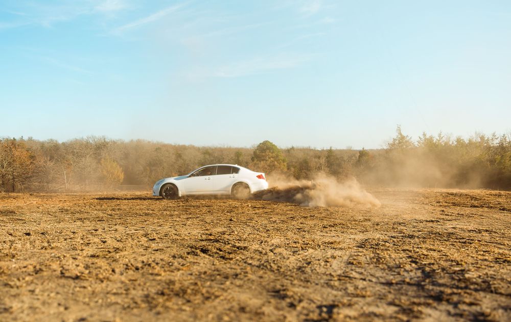 White sedan drifting on a dusty dirt field, kicking up clouds of dust with trees and blue sky in the background.