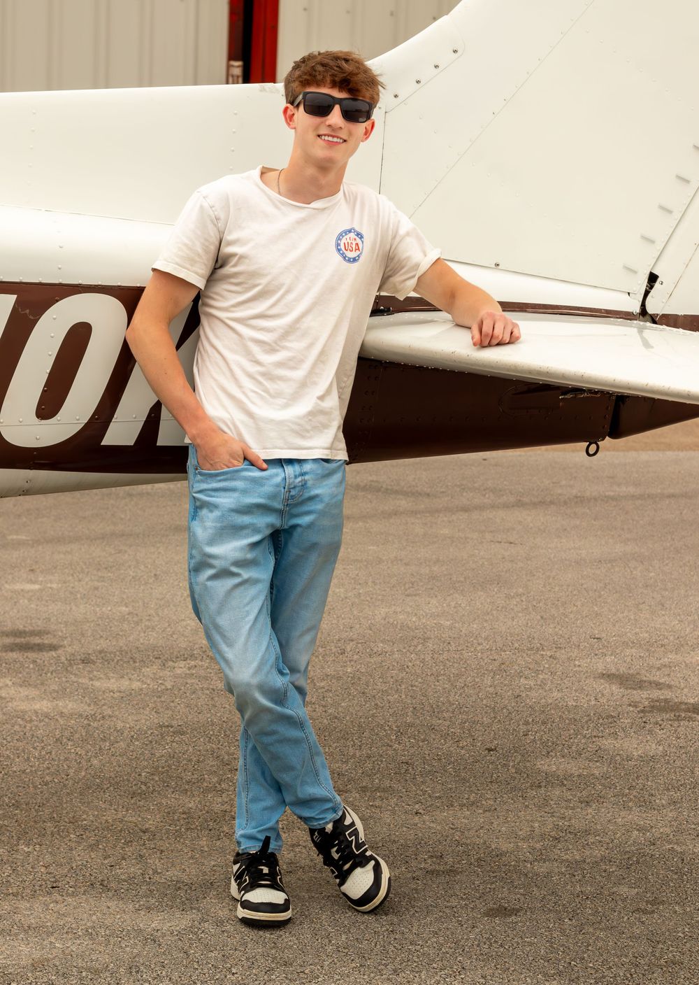 Teen boy in white t-shirt and jeans leaning against a small aircraft on a tarmac, wearing sunglasses.