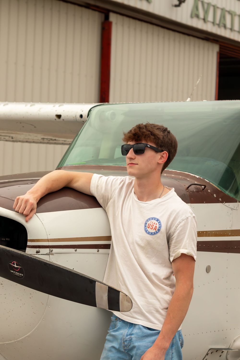 Young man in sunglasses and USA t-shirt leaning on a small aircraft at an aviation hangar.