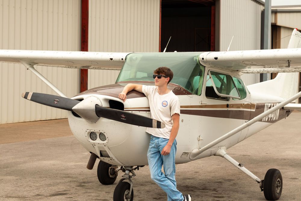 Young aviator leaning against a small Cessna aircraft in front of a hangar, wearing sunglasses and casual attire.