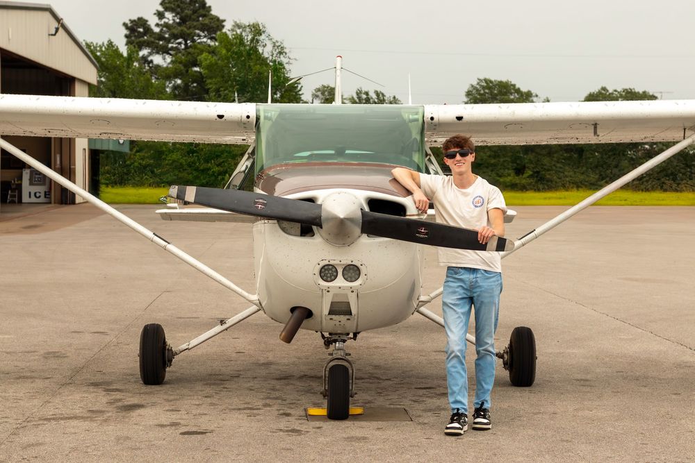 Young man posing in front of a small Cessna aircraft on a tarmac airfield, wearing jeans and sneakers.