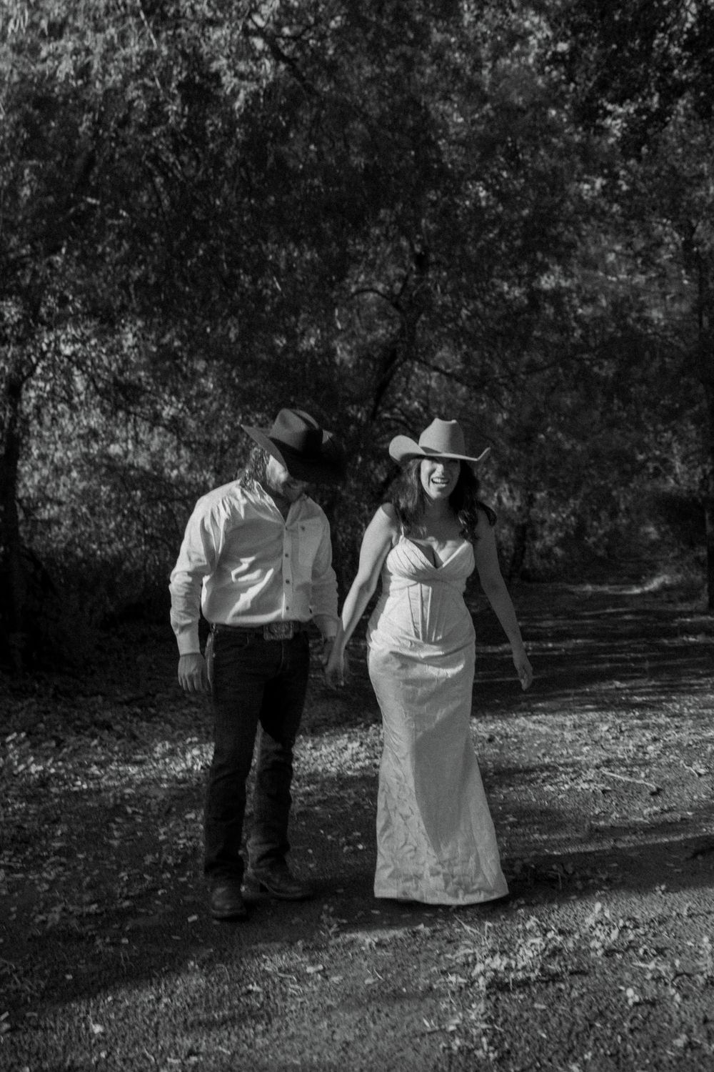 Black and white photo of a couple in cowboy hats, woman in white dress, walking on a tree-lined path.