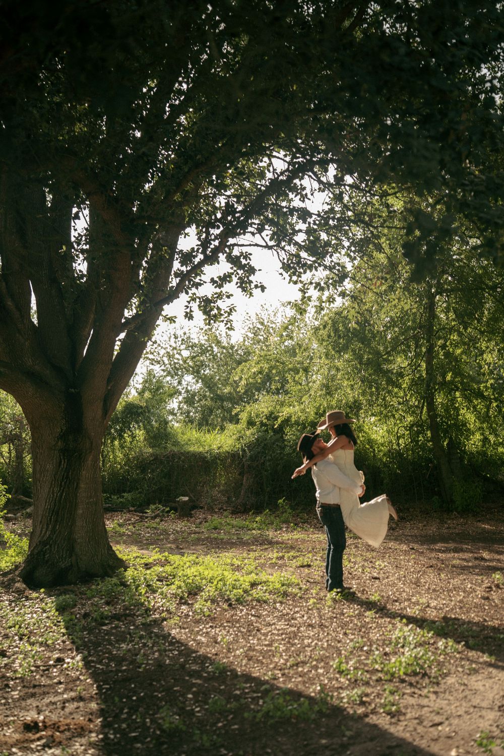 Groom lifting bride in a sun-dappled forest clearing, surrounded by lush green trees on their wedding day.