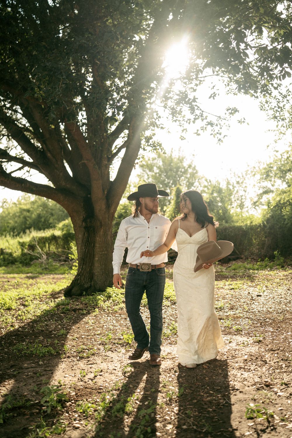 Bride in white gown and groom in cowboy hat walk together under a sunlit tree in an outdoor wedding setting.