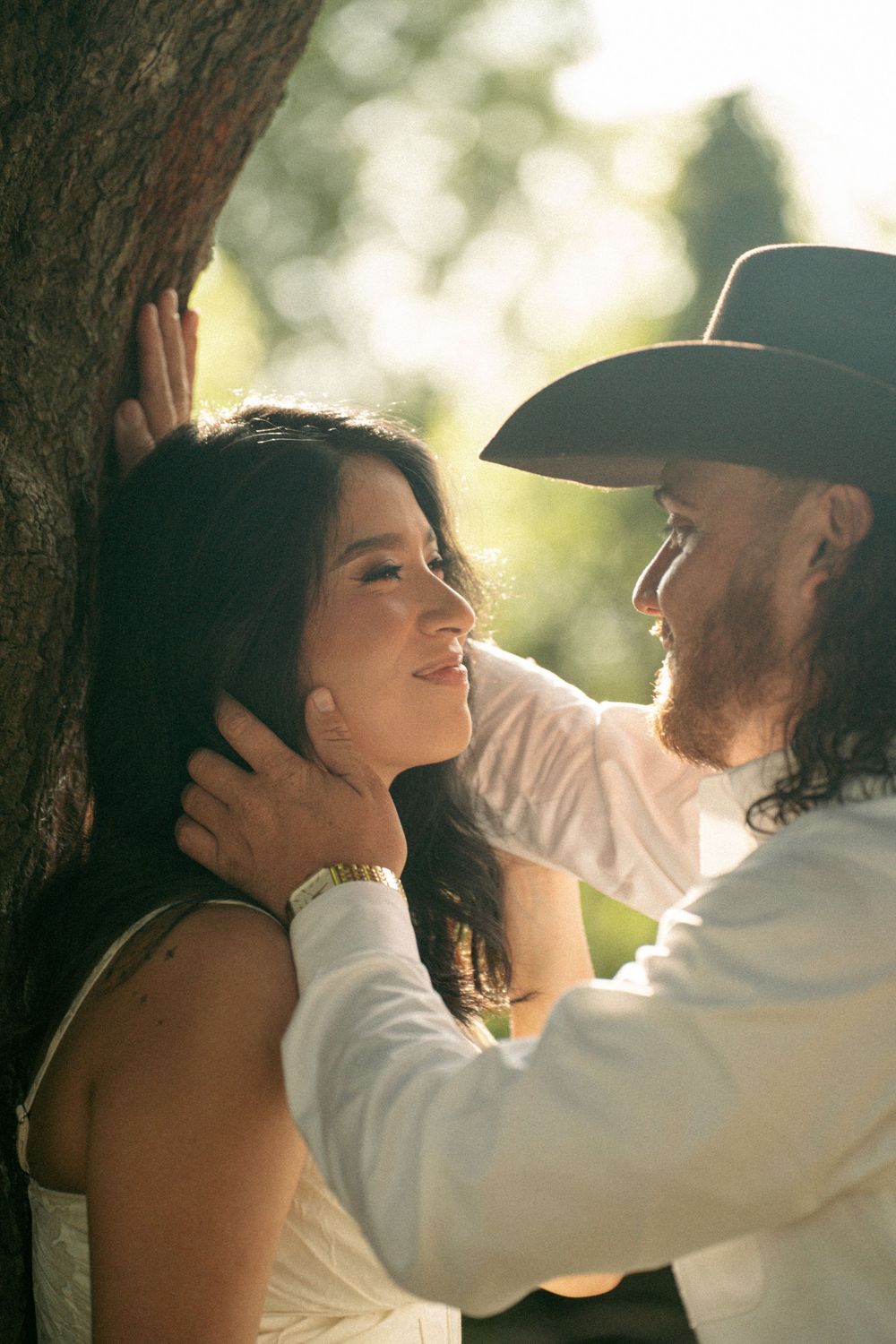 A bearded man in a cowboy hat tenderly holds a smiling woman's face near a tree in warm sunlight.