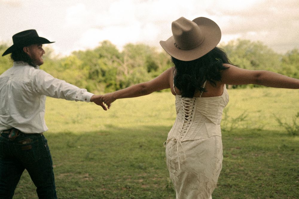 A couple in cowboy hats dancing in a green field, woman in white lace dress with arms outstretched.