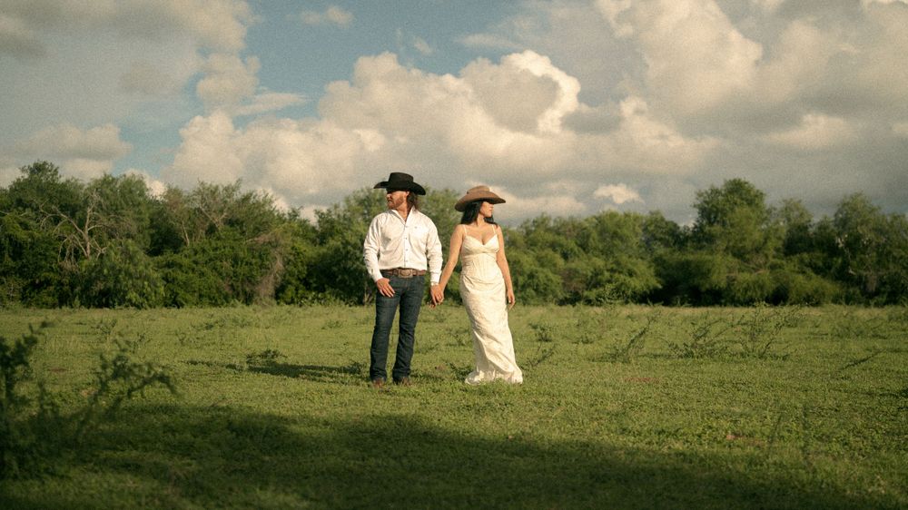 Couple in cowboy hats holding hands walking through green field with dramatic cloudy sky backdrop.