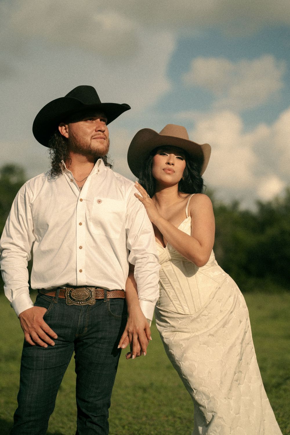 A couple in cowboy hats pose outdoors, man in white shirt and jeans, woman in white dress, green field behind them.