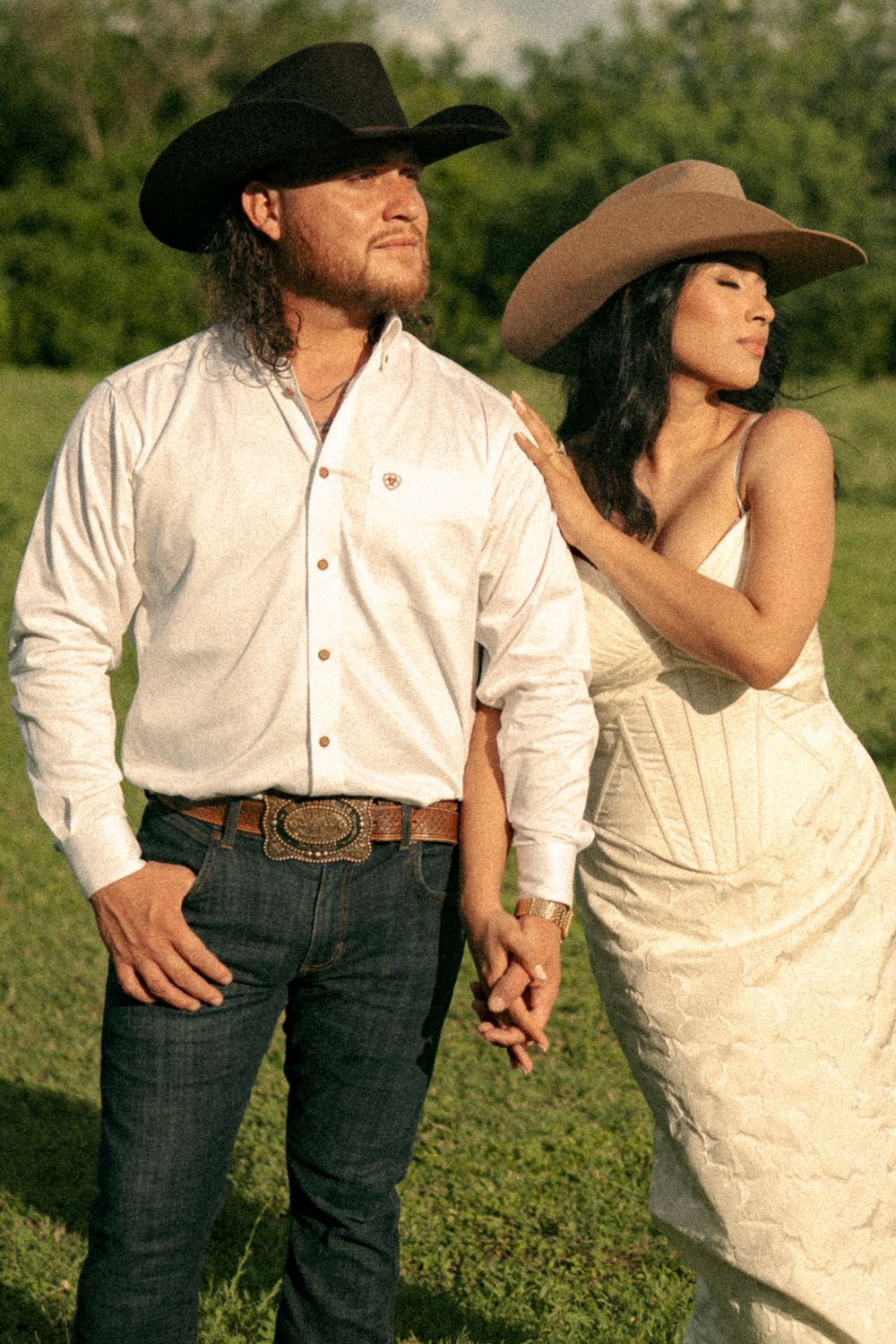 A couple in Western attire, man in white shirt and cowboy hat, woman in white dress and tan hat, posing outdoors.
