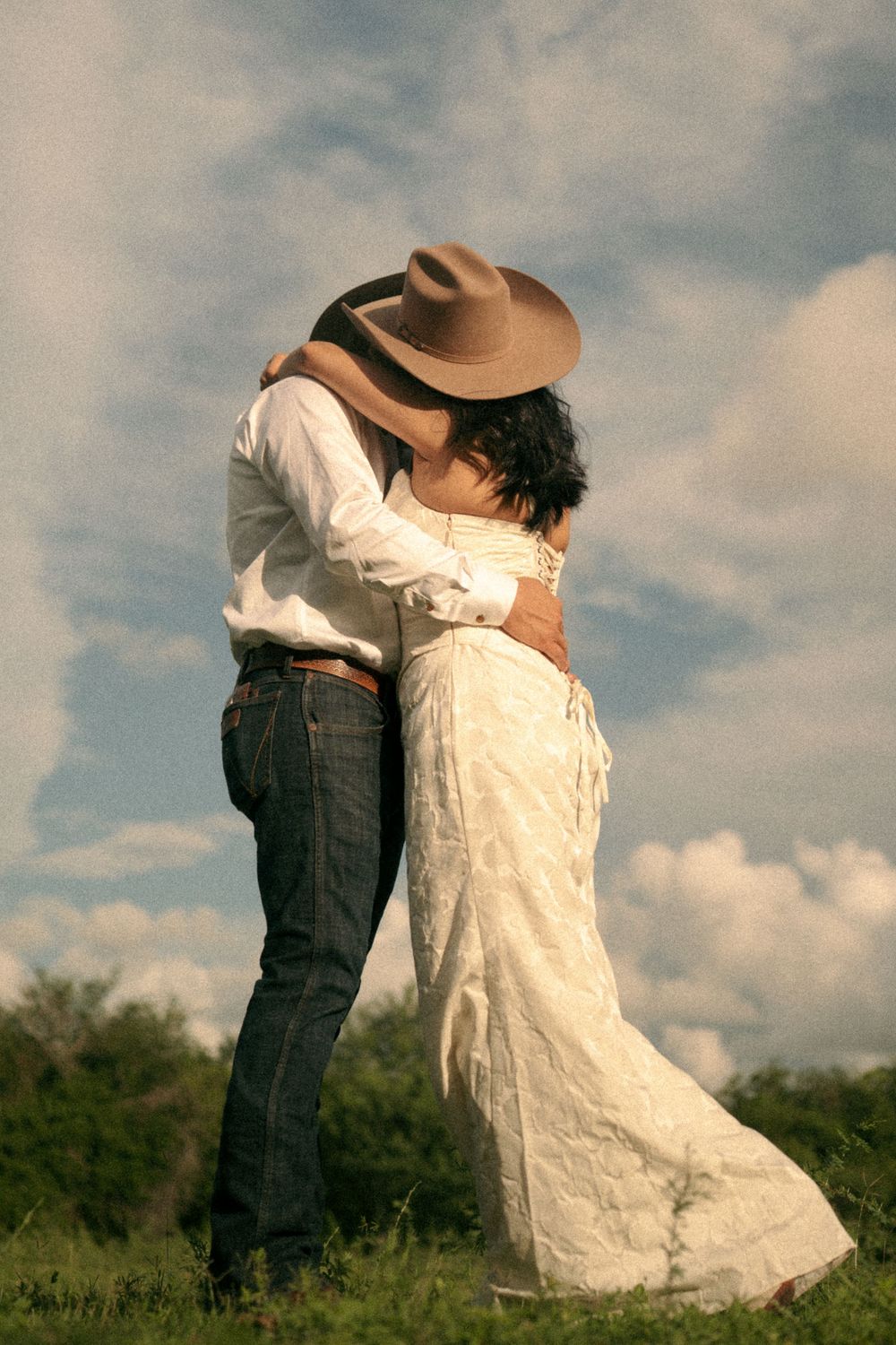 A couple in Western attire sharing a romantic embrace outdoors, the woman in a white dress and man in jeans and cowboy hat.