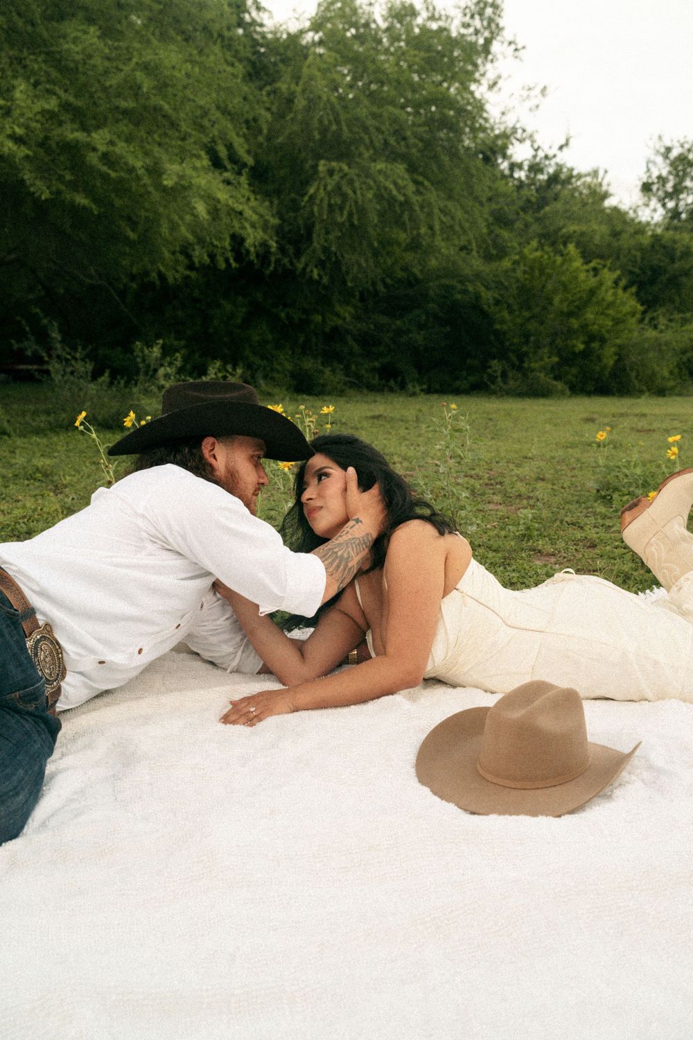 Couple in cowboy hats lying on white blanket in green field, sharing romantic moment during outdoor photoshoot.