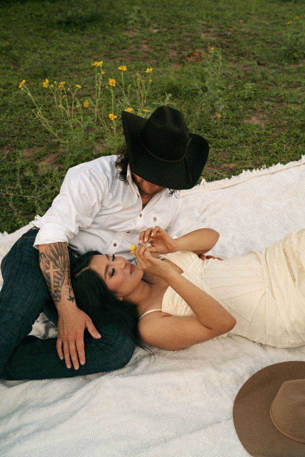 Couple relaxing on white blanket outdoors, man in black cowboy hat and woman in cream dress sharing a tender moment.